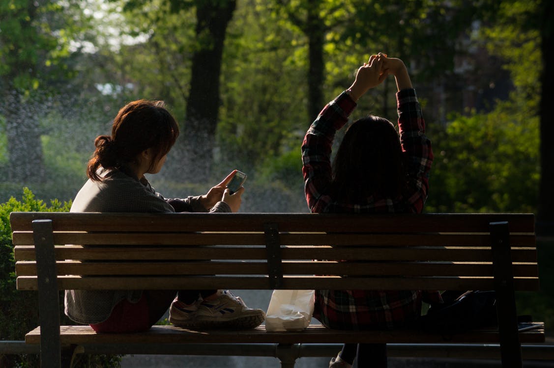 Two millennials sit on a park bench while scrolling on their phones