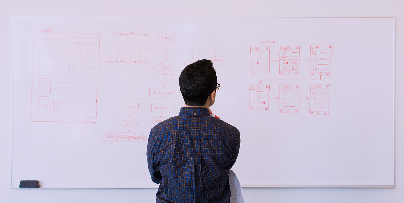 A man standing in front of a whiteboard, looking at the whiteboard with his back turned.