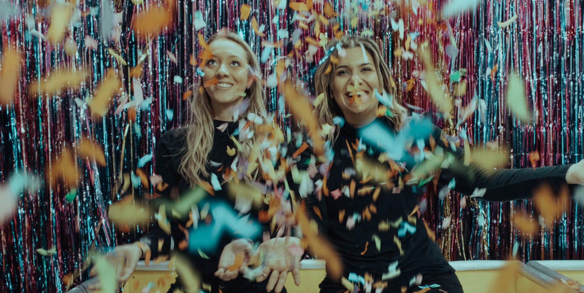 Two women smile at the camera while confetti falls on them.
