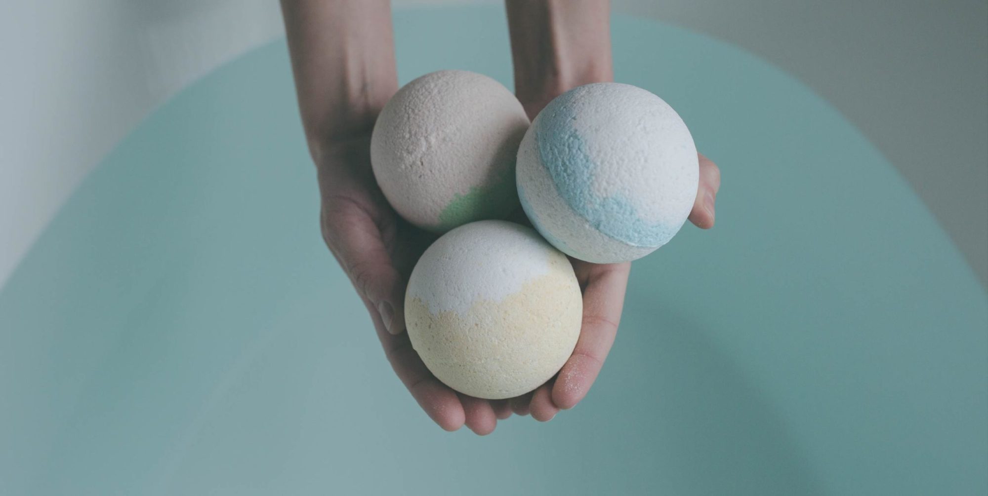 A person holding three bath bombs over a full tub of water.