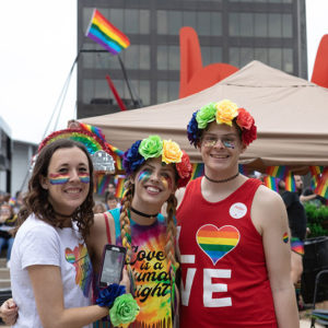 Three people smiling and standing close together wearing rainbow colored clothing to celebrate Pride.