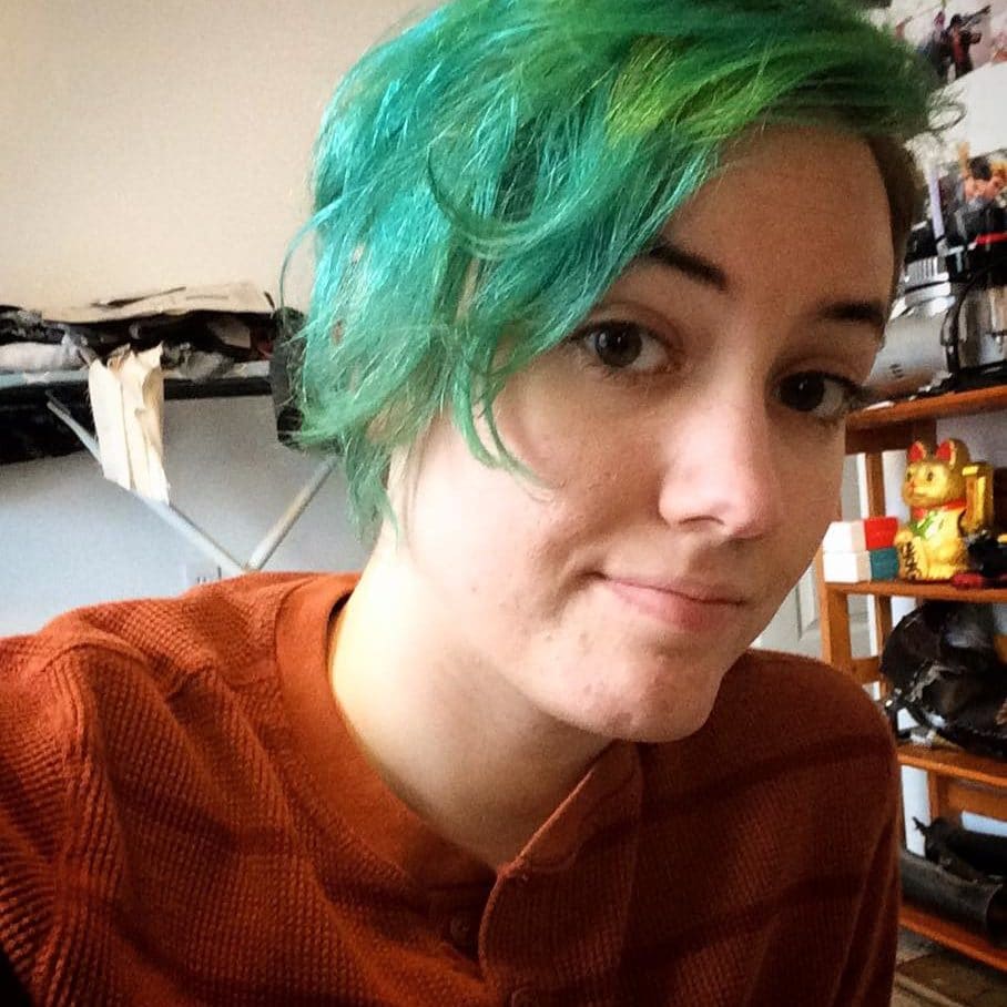 Ro looks at the camera with bright green hair, red shirt, and a small smile..